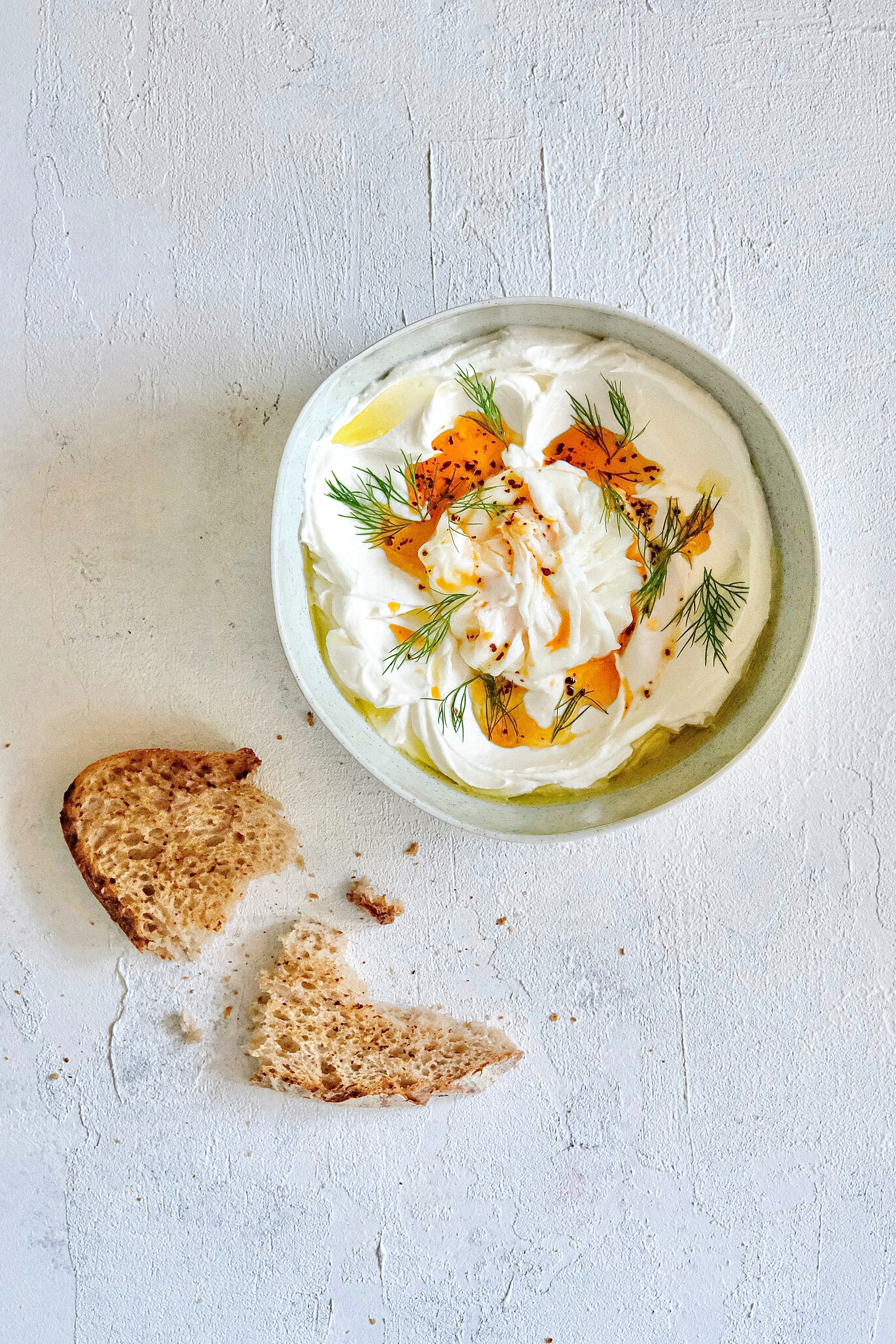 A bowl of a yogurt-dip with pieces of fresh dill, spices and oil visible on the surface. Next to the bowl is a small piece of toasted brown bread, broken in half.