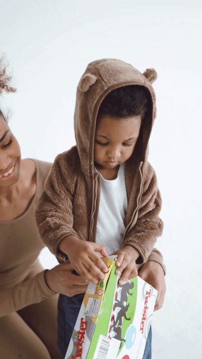 A toddler in a hooded sweatshirt with two small bear ears on the hood is standing, trying to open the board game box he is holding. An adult kneels behind him, helping to support him.